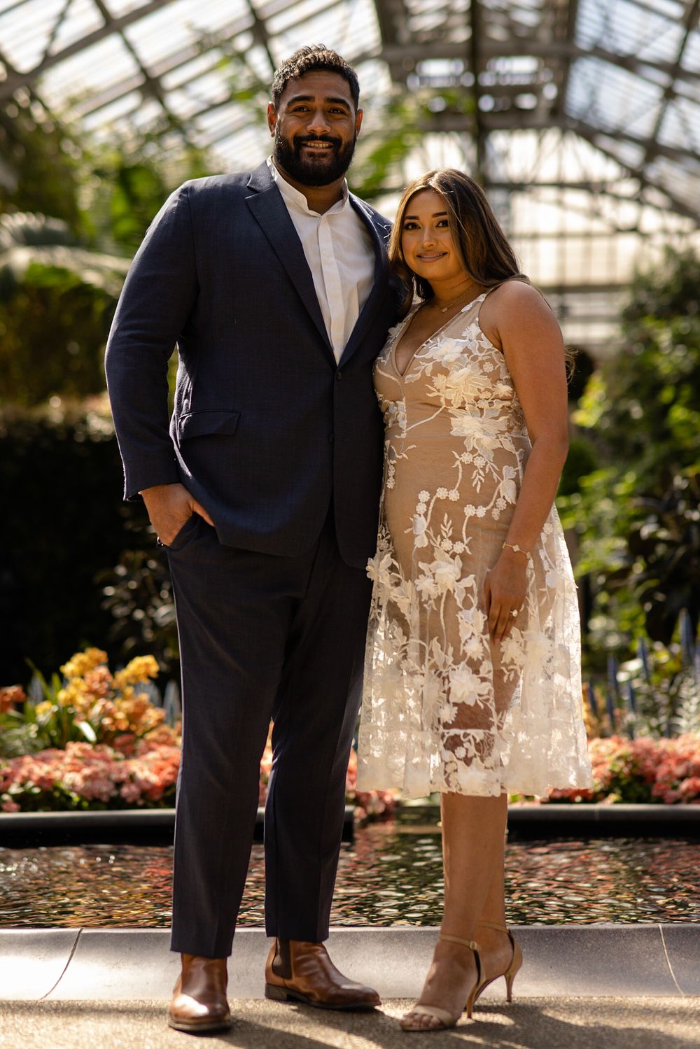 Couple in formal wear poses in a greenhouse with blooming flowers in the background.