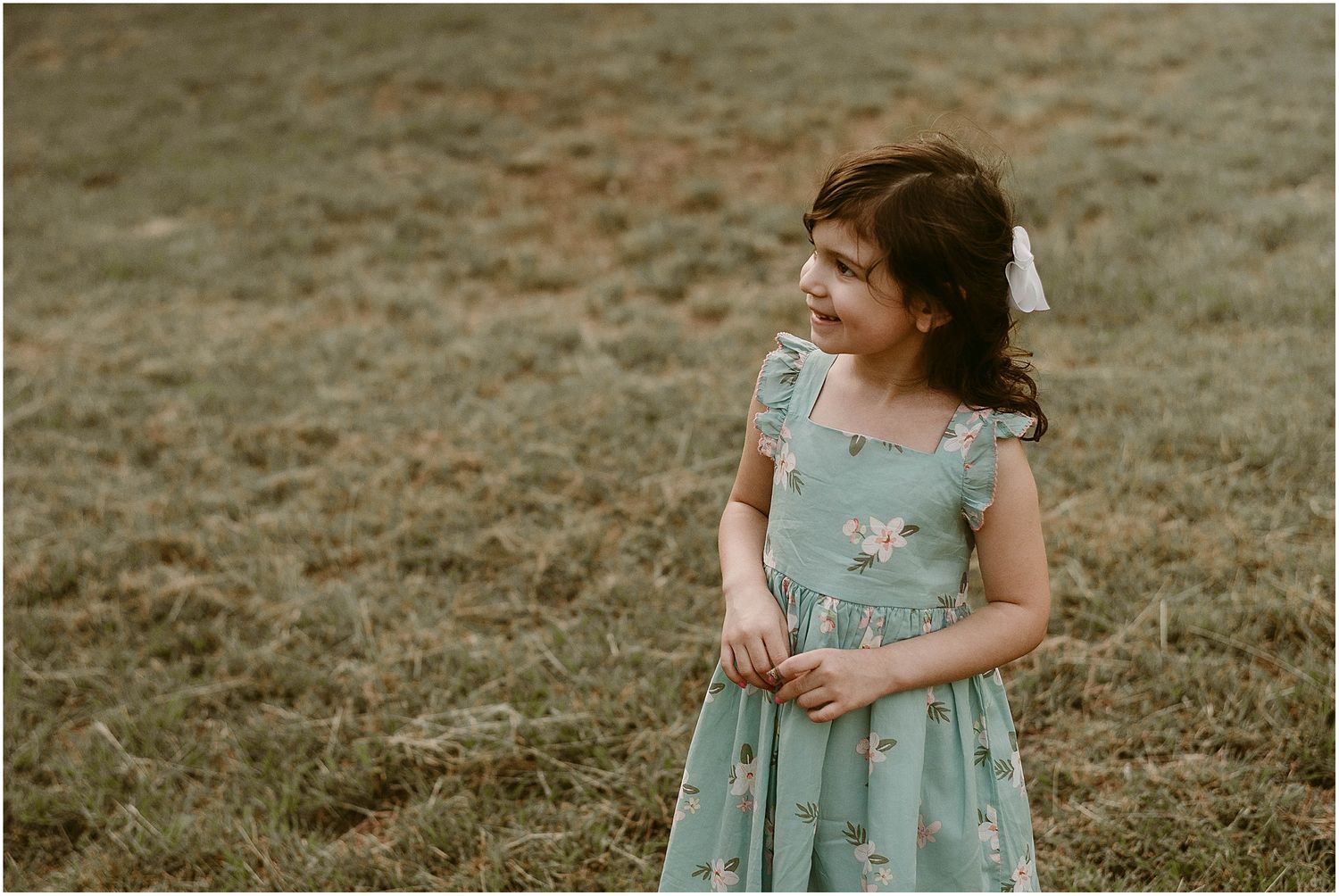 Young child in floral mint dress stands in grassy field during golden hour.