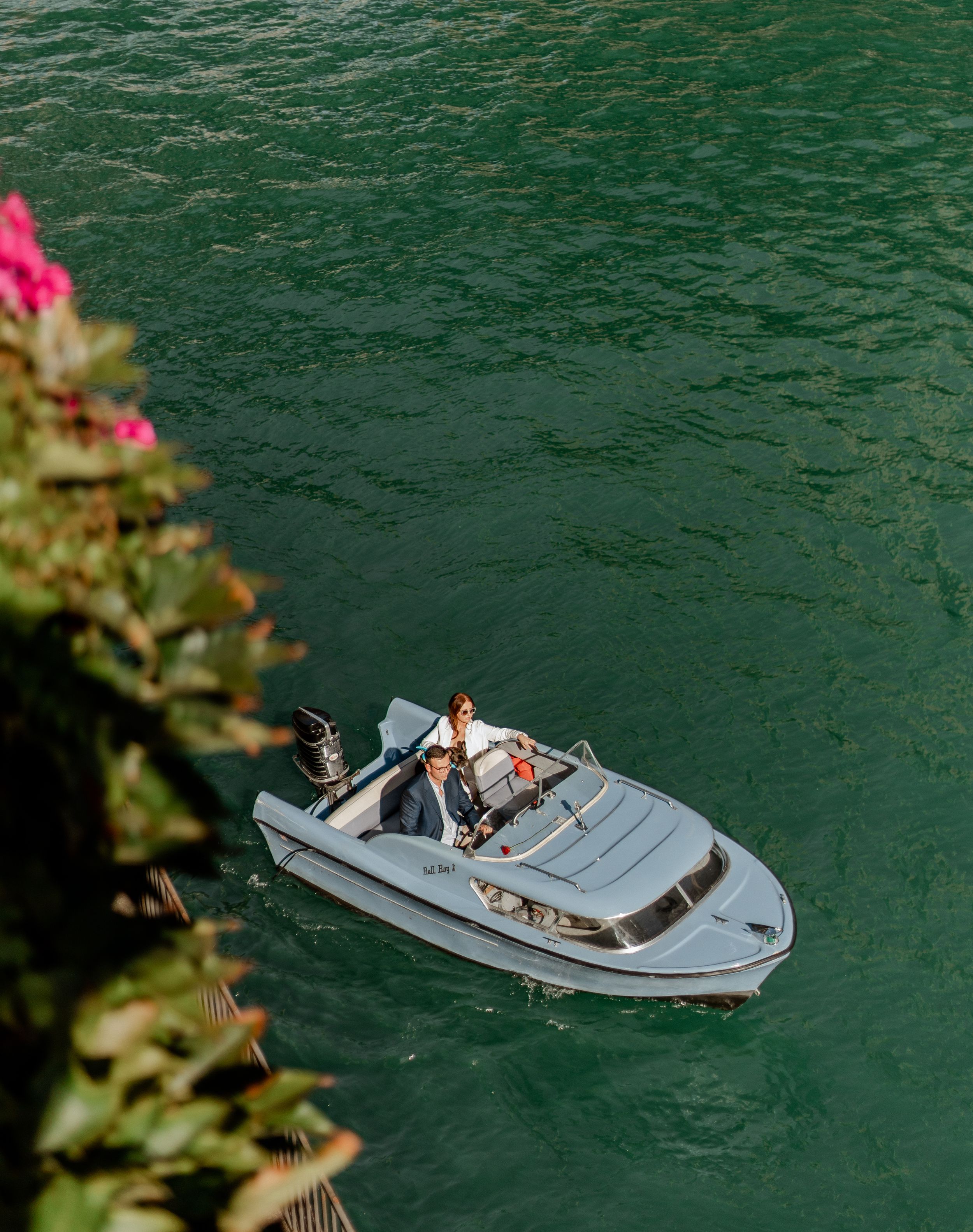 Aerial view of small white boat cruising on emerald green water with pink flowers in foreground.