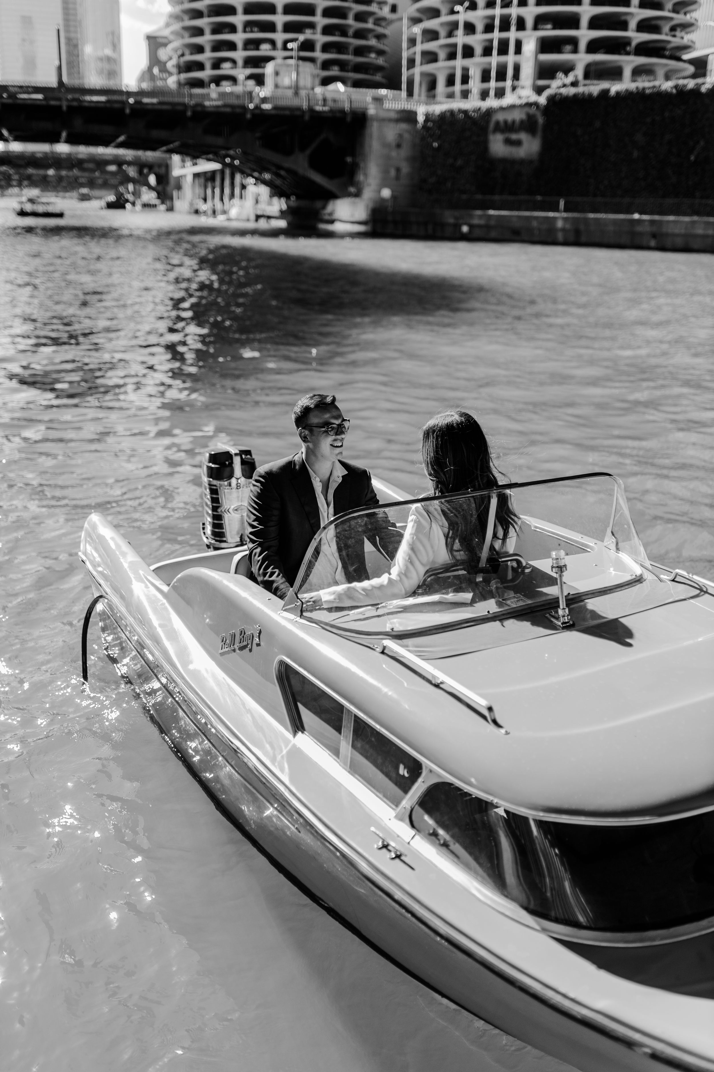 Black and white photo of couple on vintage boat ride along urban riverfront.
