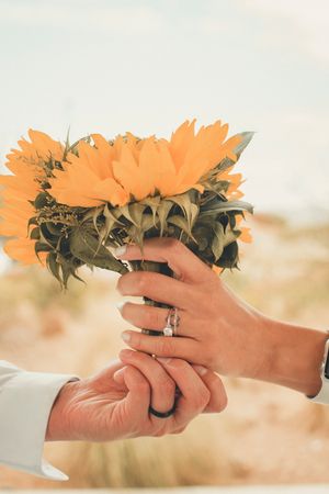 Hands holding a bouquet of sunflowers with a wedding ring visible in romantic outdoor lighting.