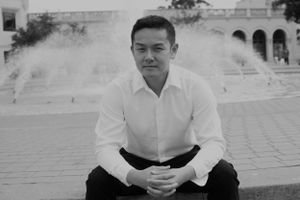 Black and white portrait of person sitting outdoors near architectural columns.