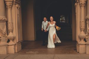Wedding couple emerging from ornate archway in classic architectural setting.