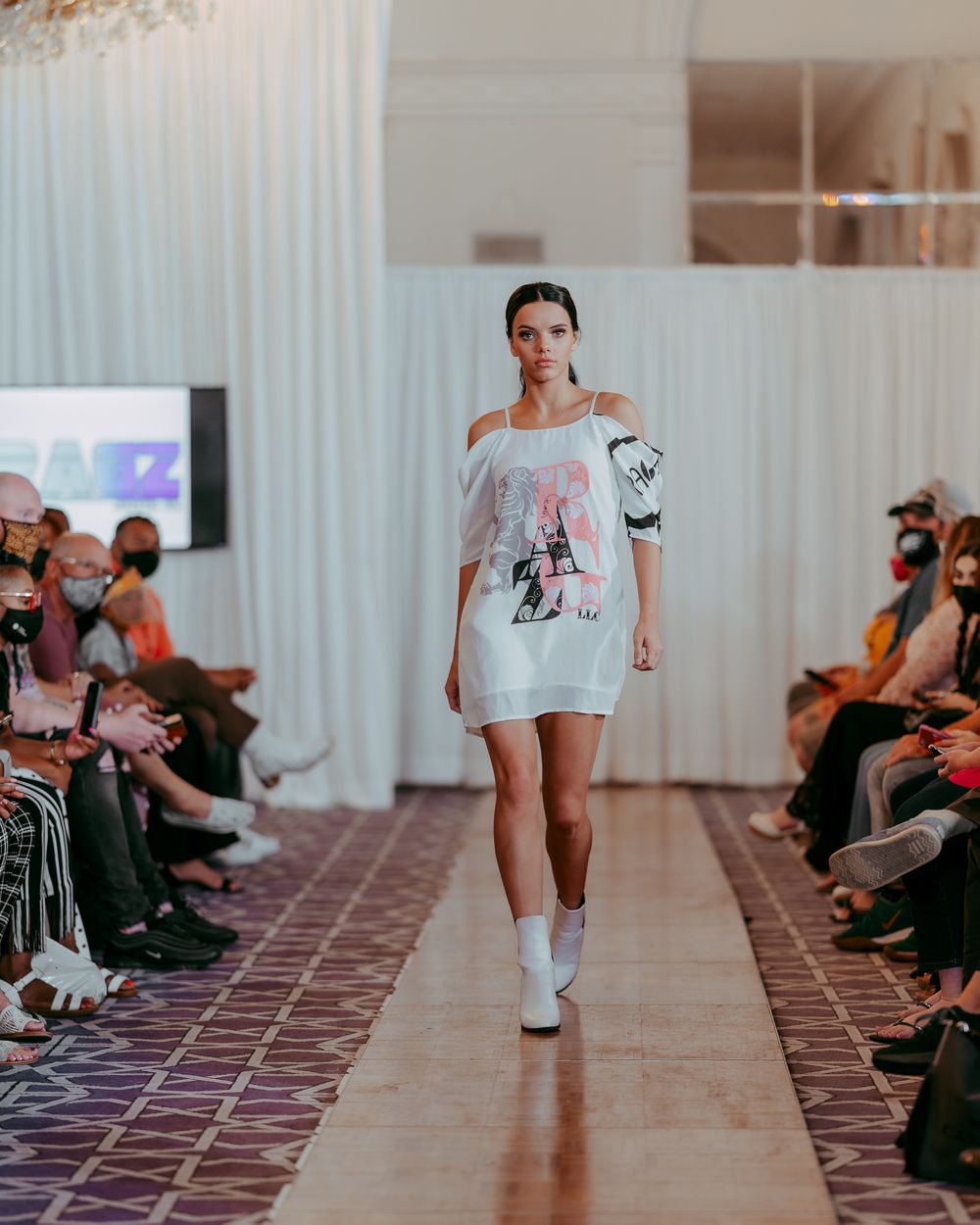 Model walks runway in white graphic t-shirt and matching white boots during fashion show.
