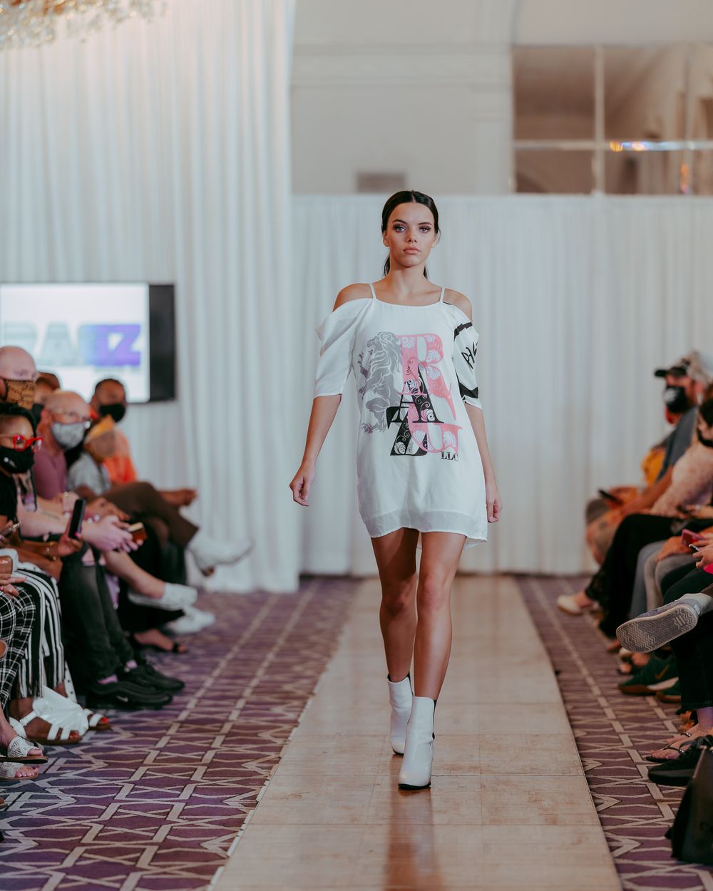 Fashion model presents casual white ensemble on pink-tiled catwalk with audience sitting in background.