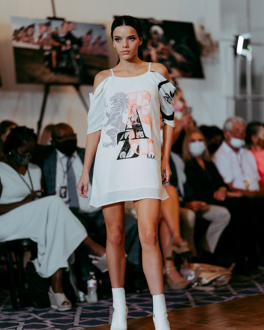 Model walks catwalk in white graphic t-shirt and matching boots against dark audience.