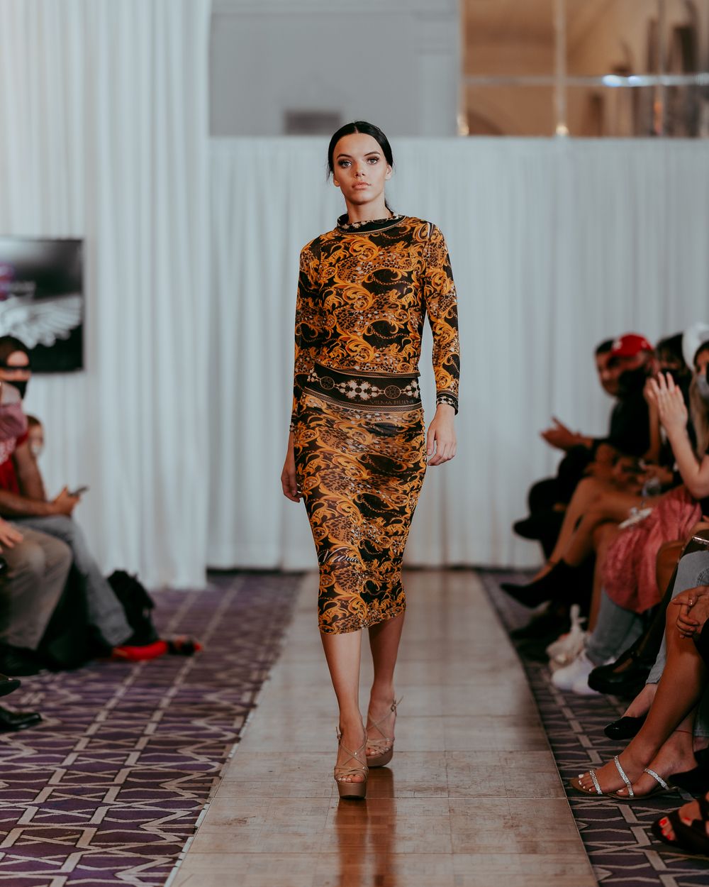 Fashion model walking runway in orange and black patterned bodycon dress during fashion show.