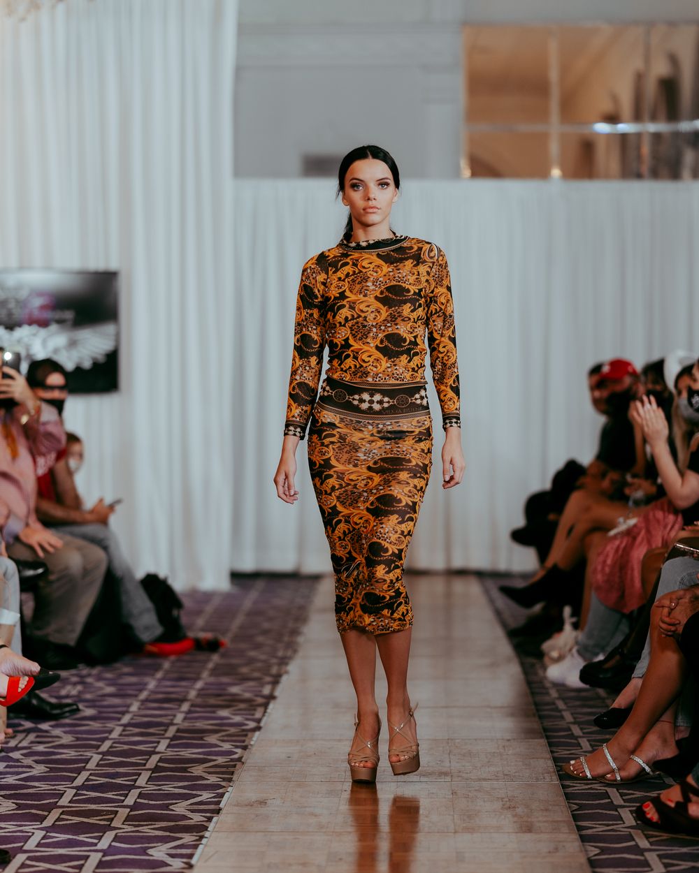 Form-fitting dress with ornate orange pattern presented at luxury fashion event.