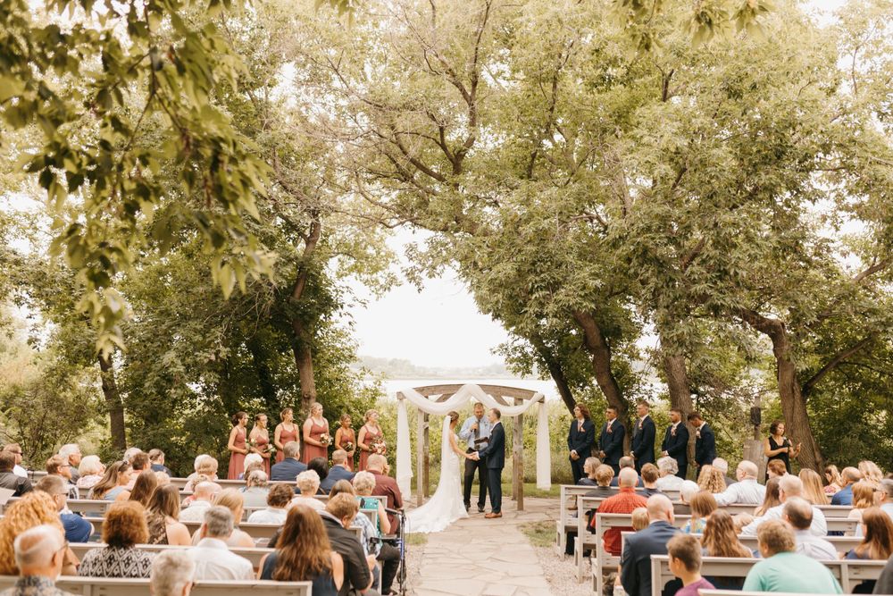 Outdoor wedding ceremony with bridesmaids in rust dresses and guests seated under lush trees.