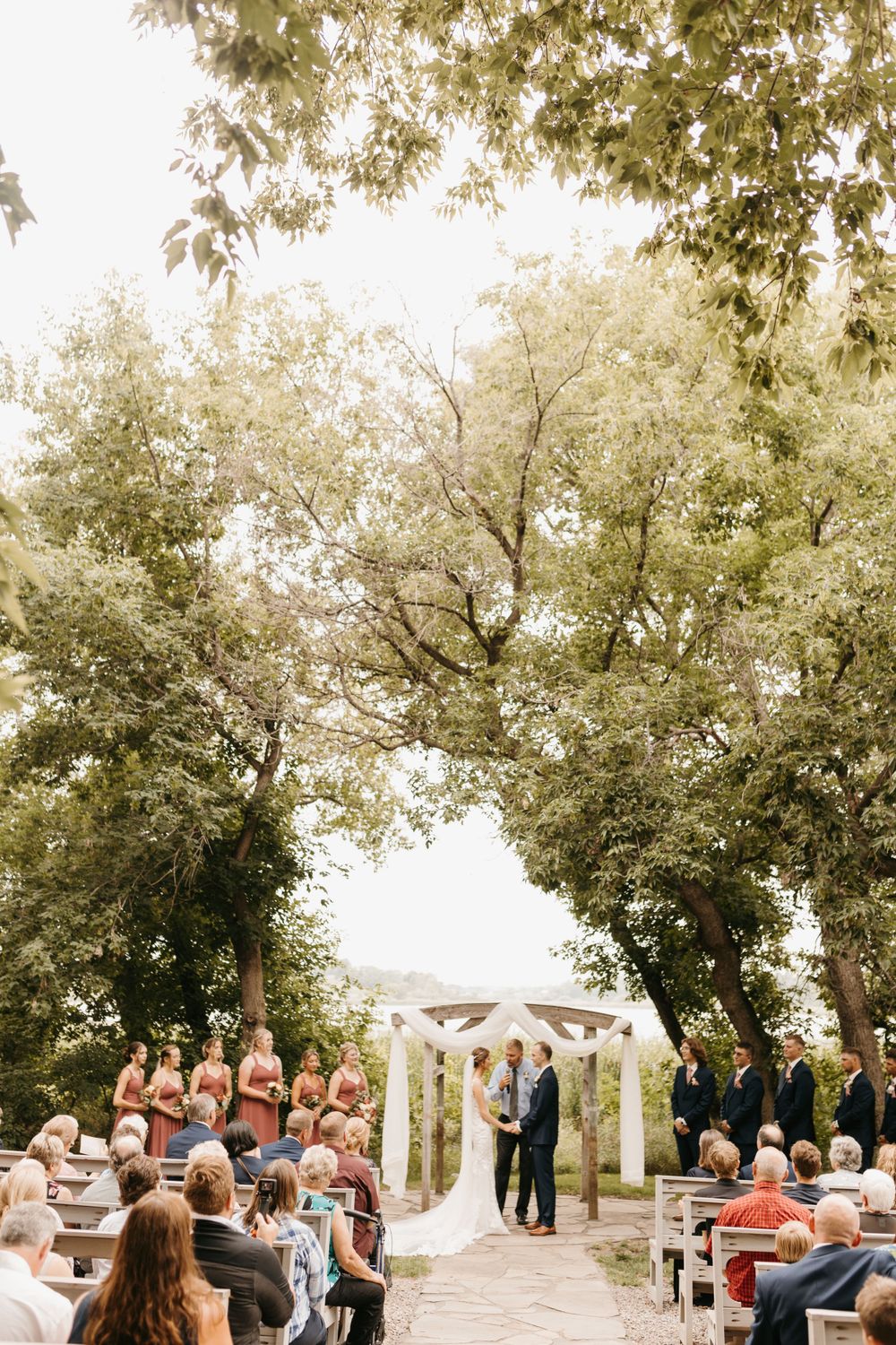 Outdoor wedding ceremony under large trees with white arbor, guests seated on wooden benches along aisle.