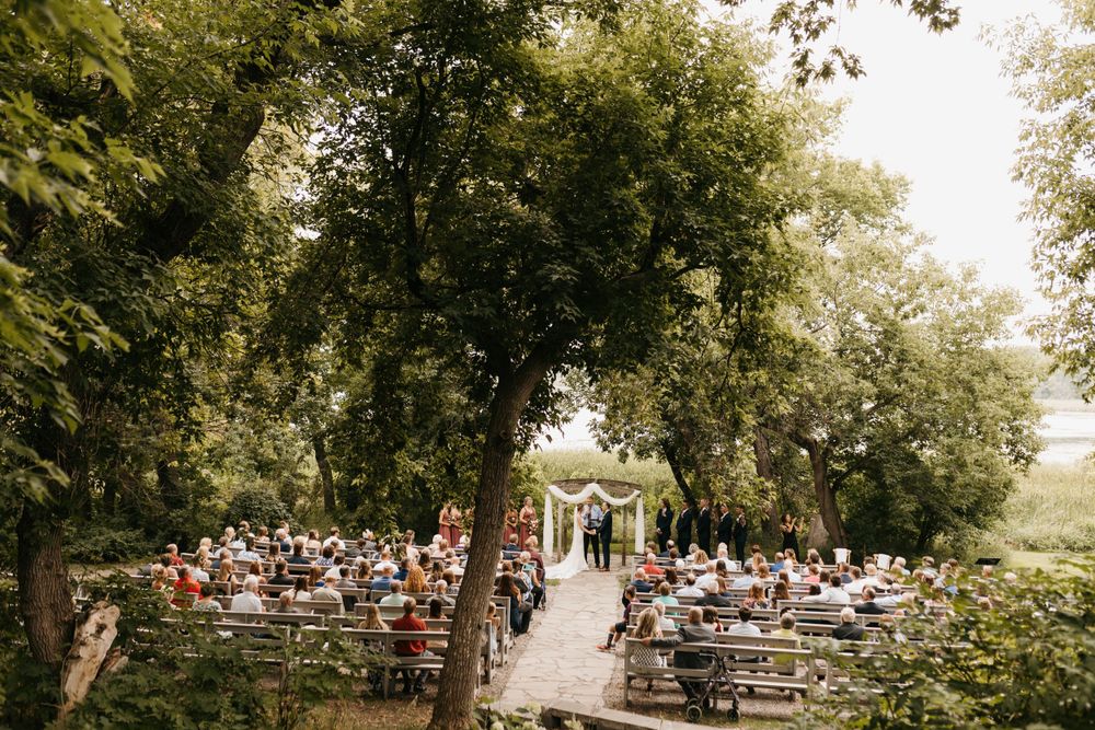 Wide aerial view of outdoor wedding ceremony with guests seated on wooden benches under trees.