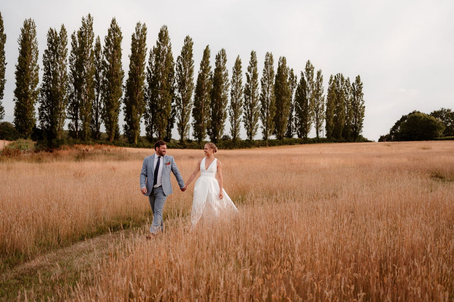 A newlywed couple walk hand in hand together through a golden grassy field at their Silchester Farm wedding