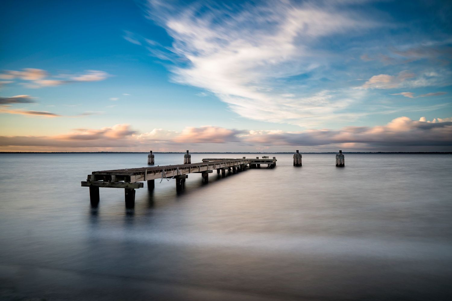 Long exposure of a weathered pier extending into calm waters under dramatic clouds at sunset.