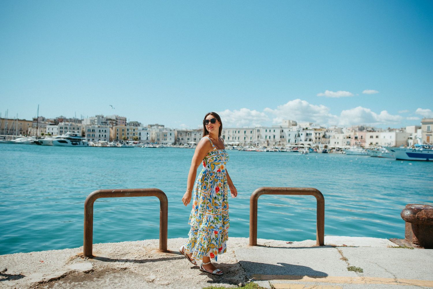 Woman in floral dress standing on pier overlooking Mediterranean harbor with boats and buildings.
