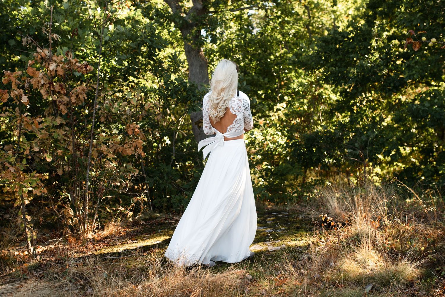 Bride in flowing white dress twirls in sunlit forest setting during wedding photoshoot.