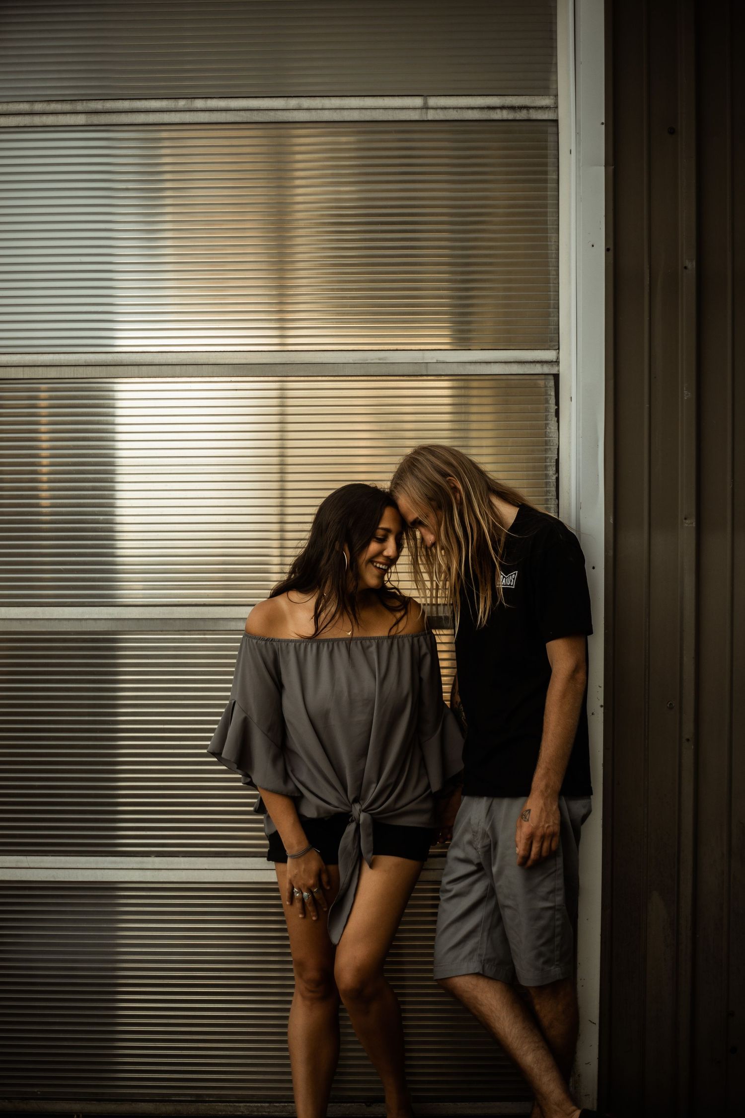 Couple sharing intimate moment against industrial window with venetian blinds.