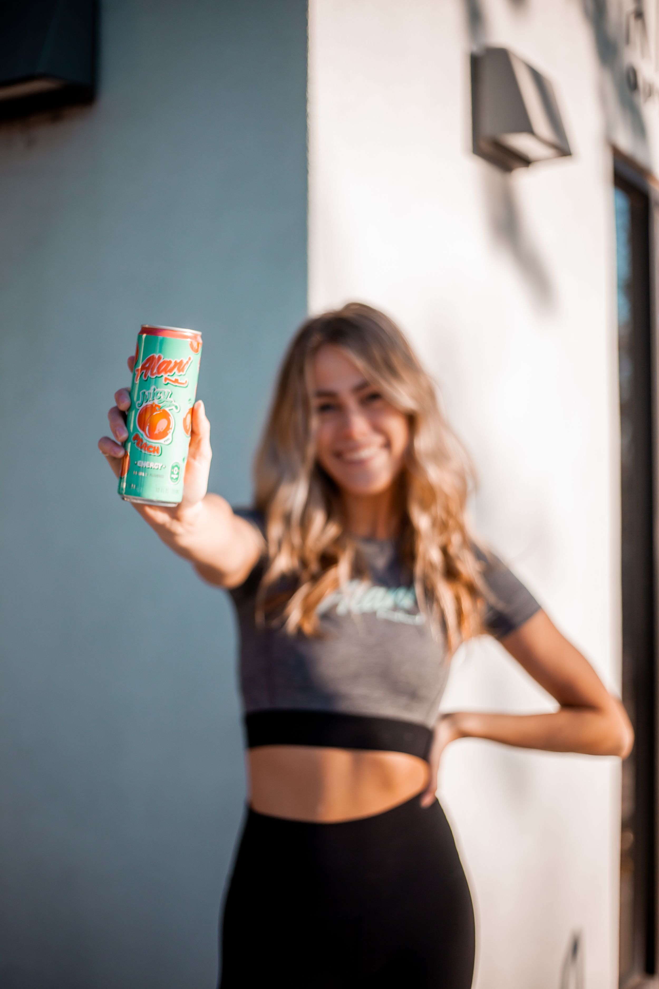 Athletic woman in workout attire holding up a teal and orange beverage can against a white wall.