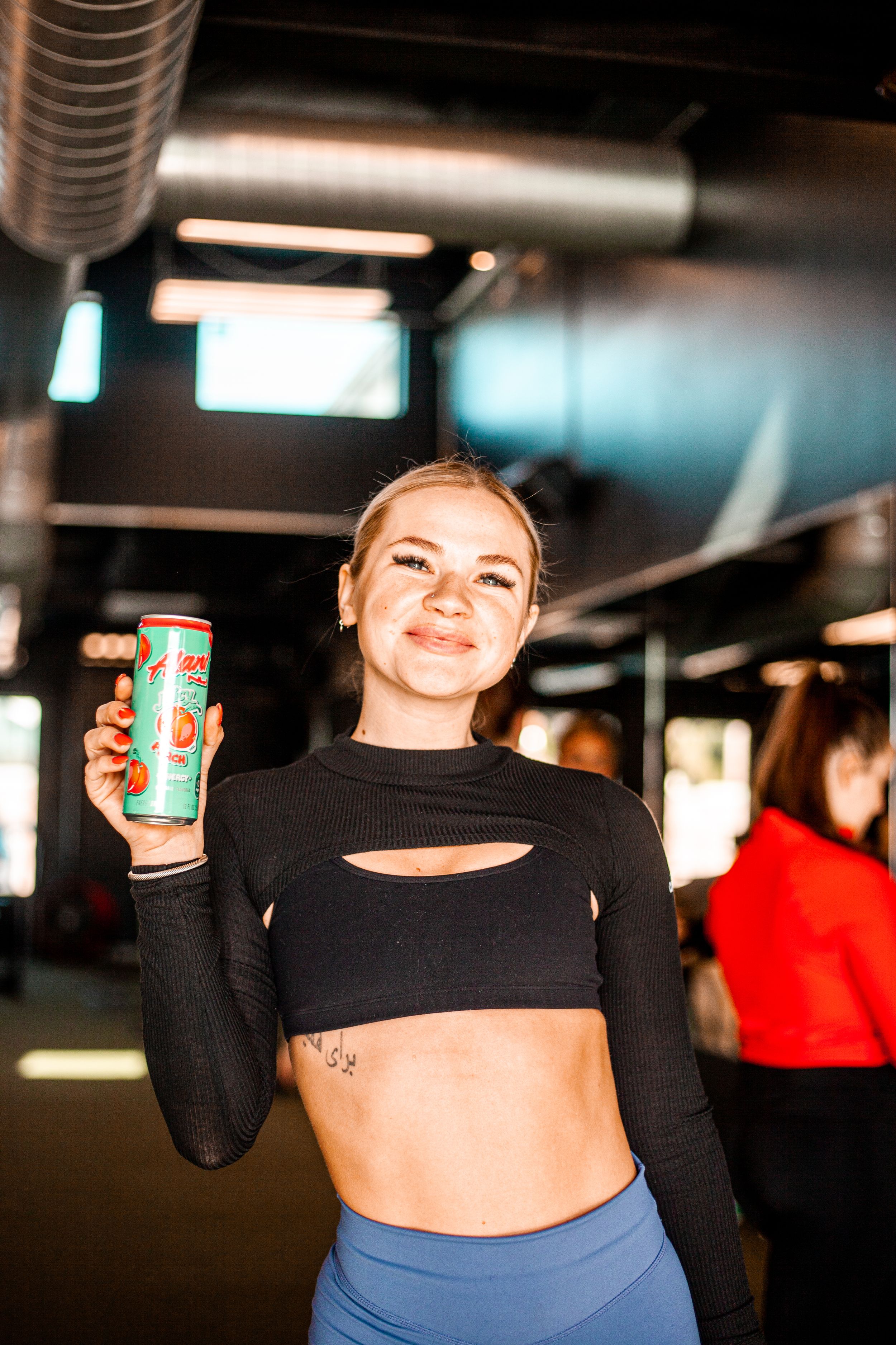 Fitness enthusiast in black crop top and blue leggings holding energy drink in dimly lit gym.