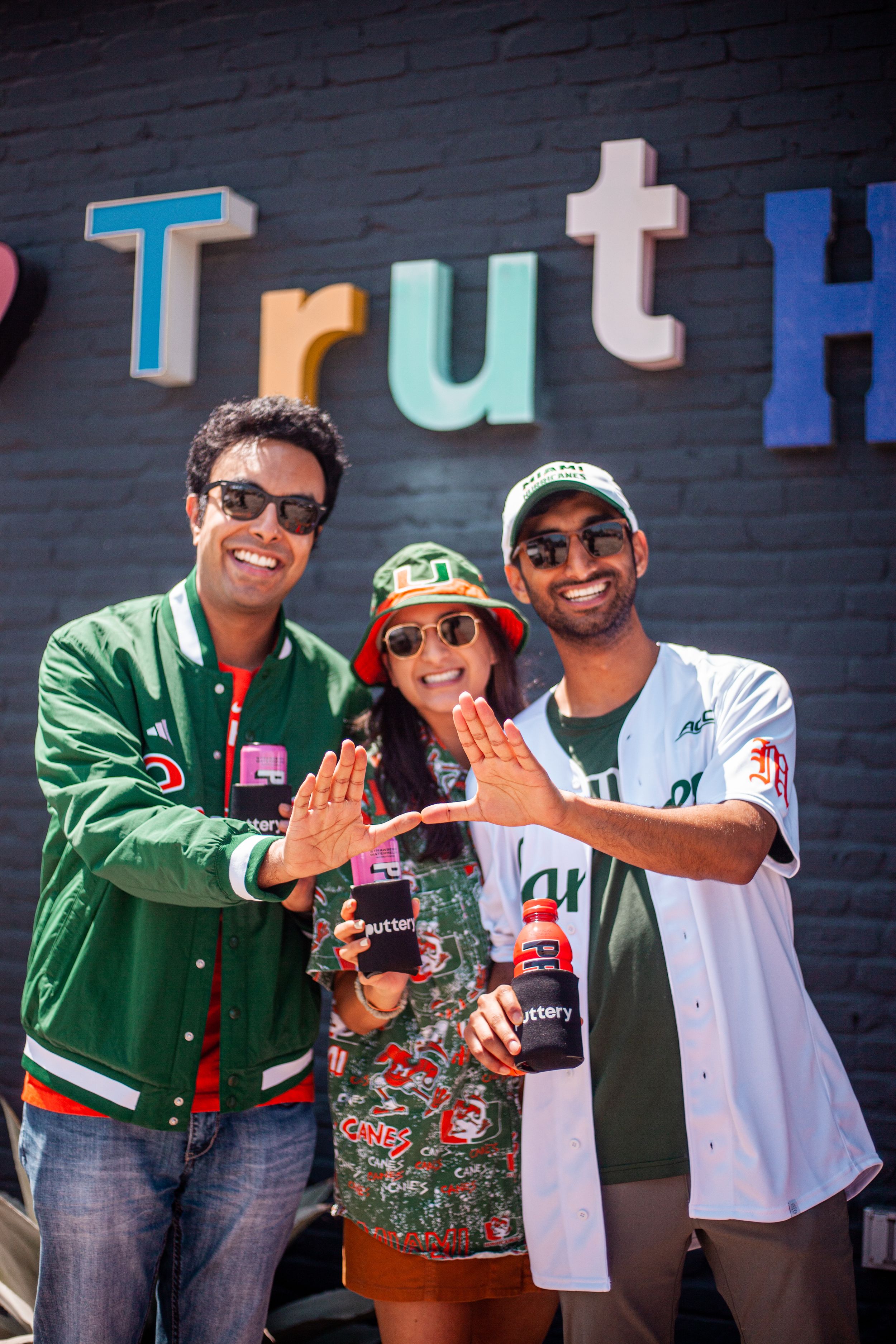 Three people posing with beverages in front of colorful Truth wall sign.