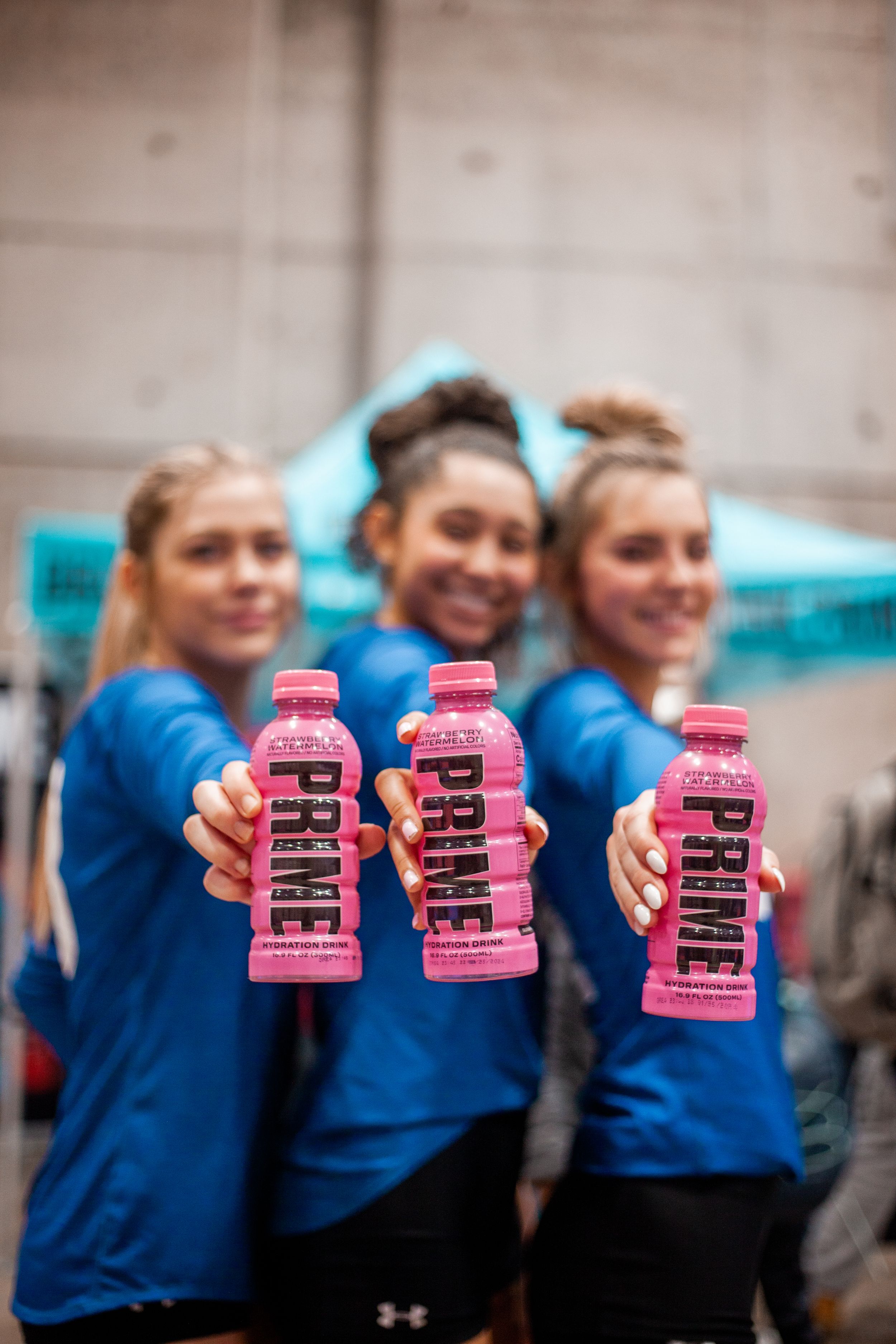 Athletes in blue uniforms holding pink Prime energy drink bottles against concrete wall.