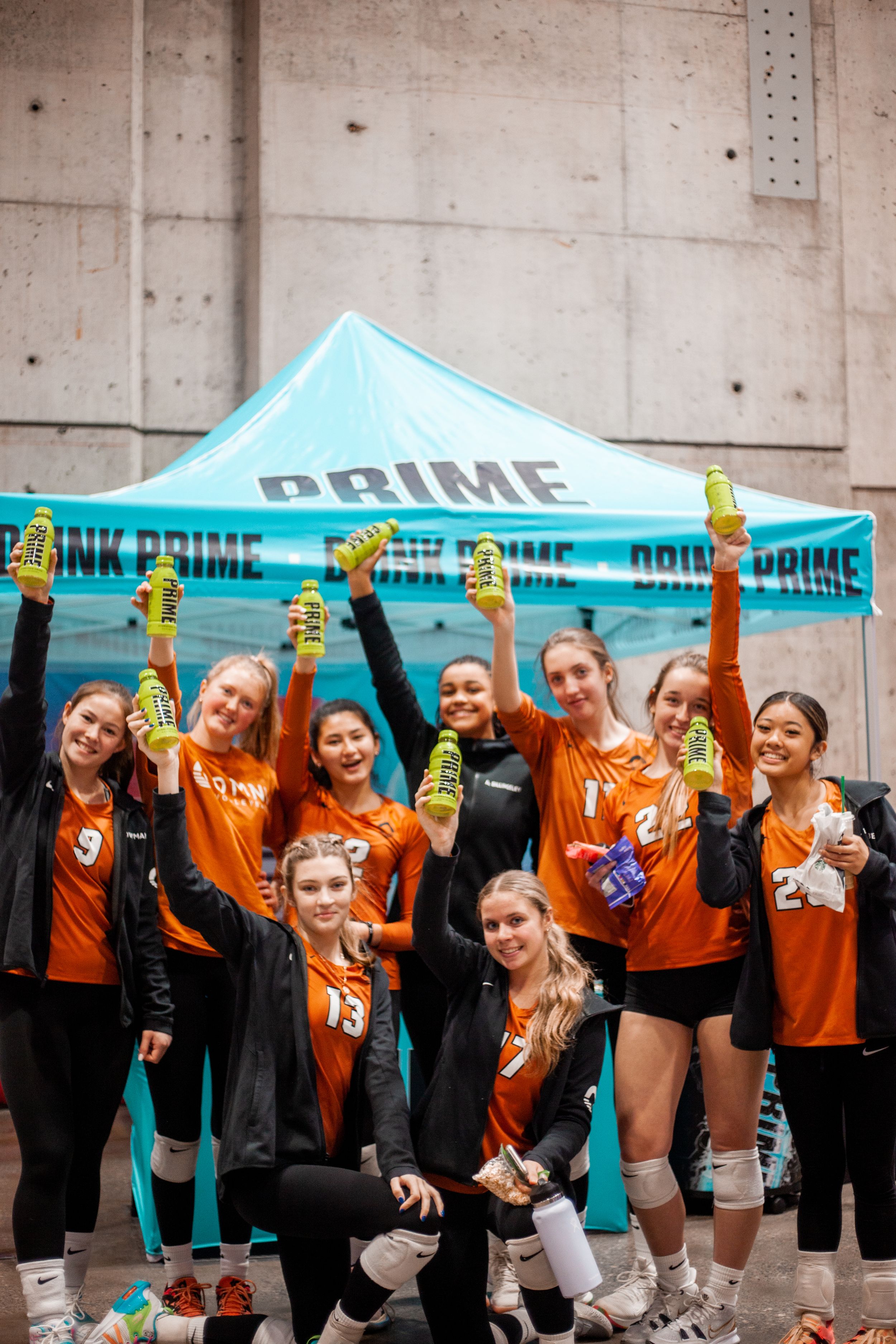 Group of volleyball players in orange jerseys celebrating under a blue tent with Prime branding.