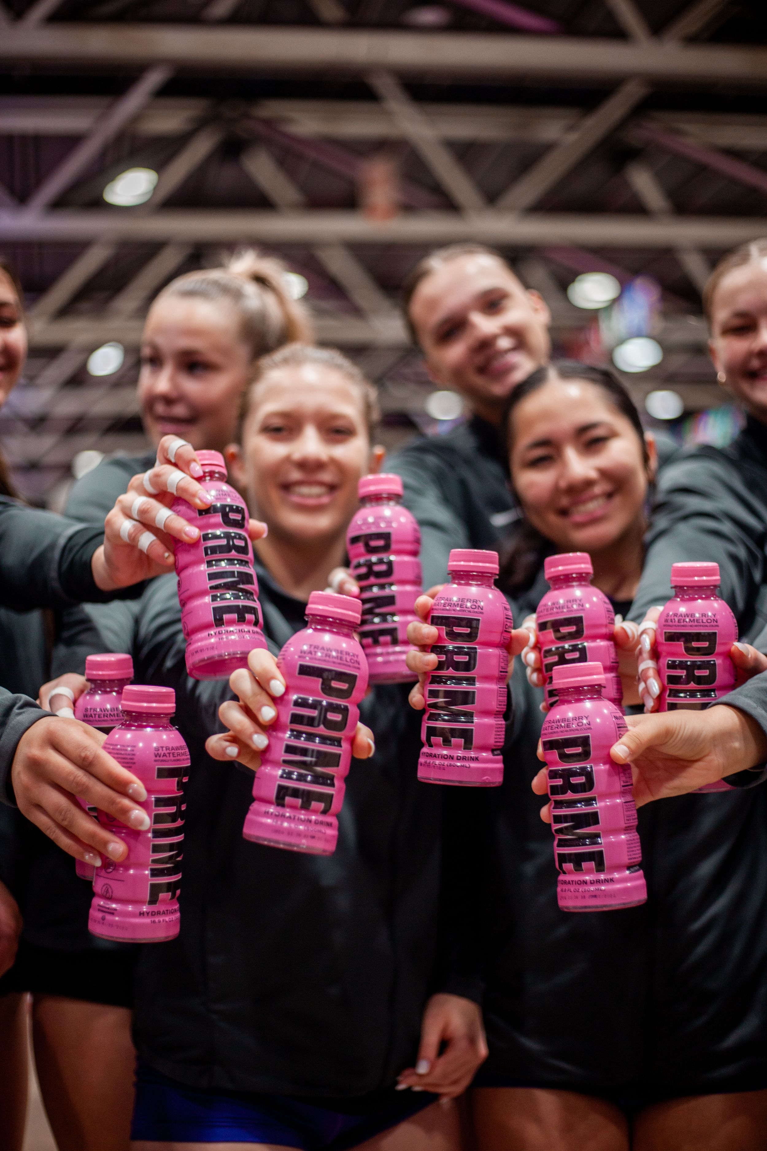 Athletes in dark uniforms holding up pink Prime energy drinks in an indoor sports venue.