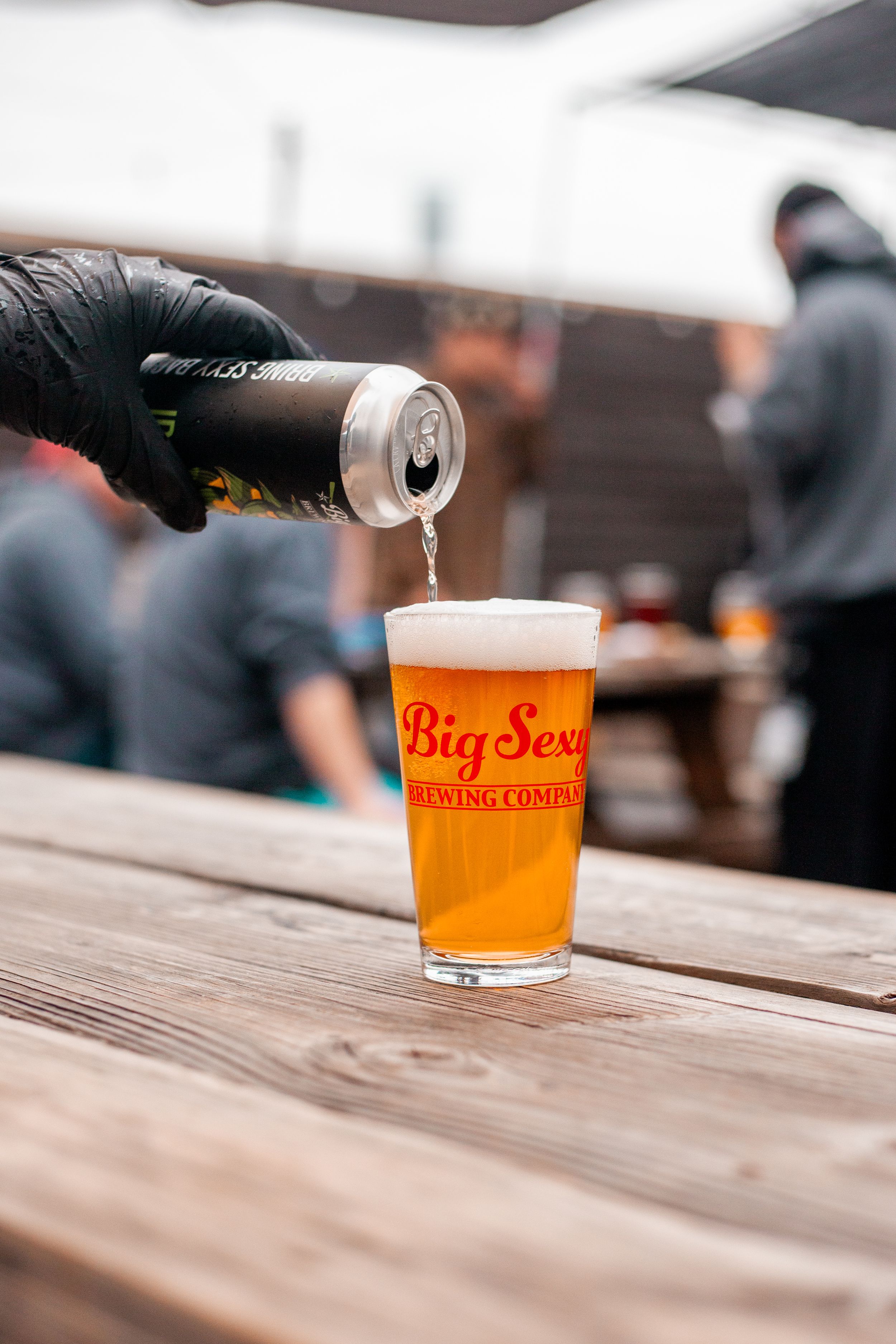 Hand in black glove pouring beer from can into Big Sexy Brewing Company branded glass on wooden table.