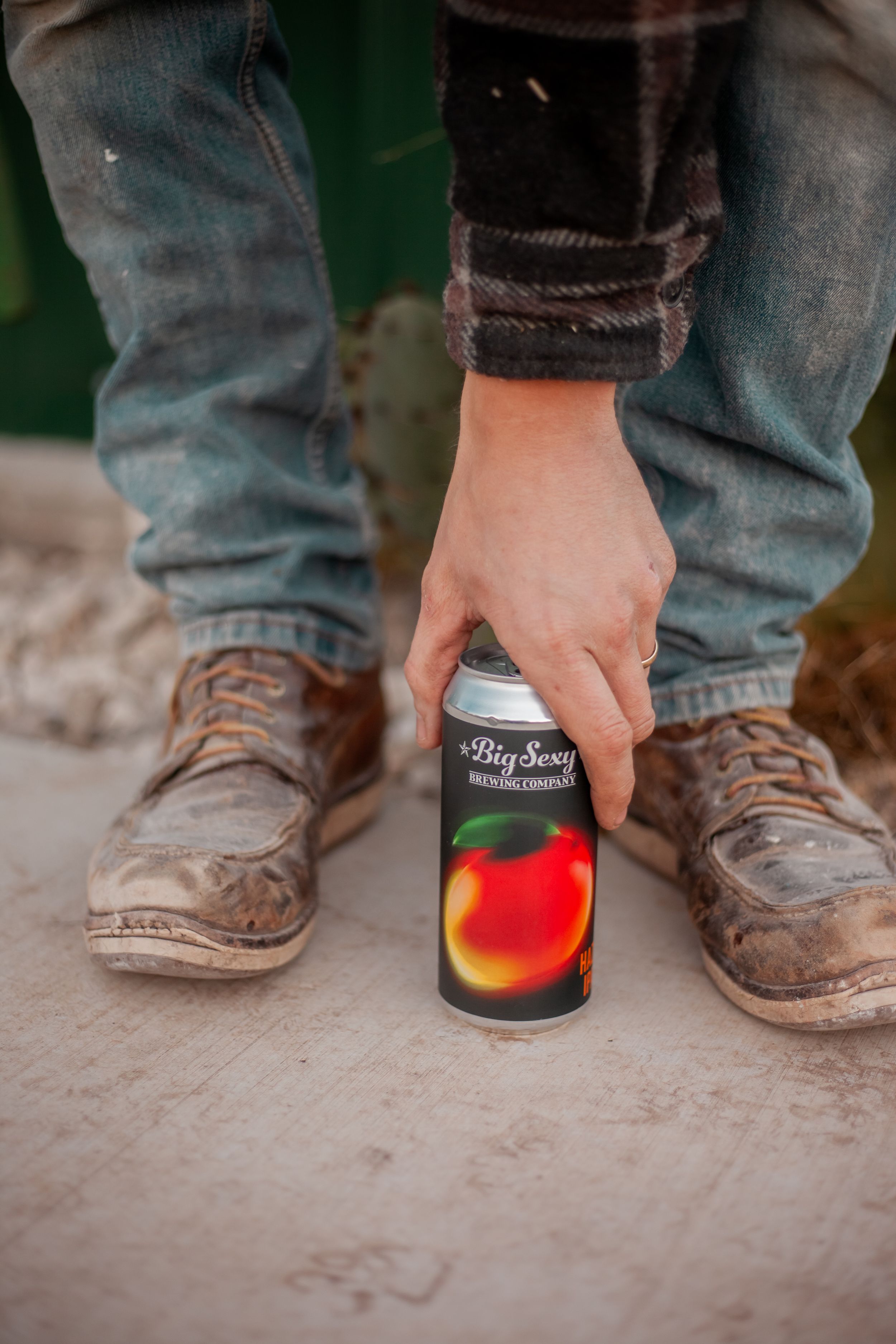Close-up of worn boots and denim jeans with person reaching for beverage can.