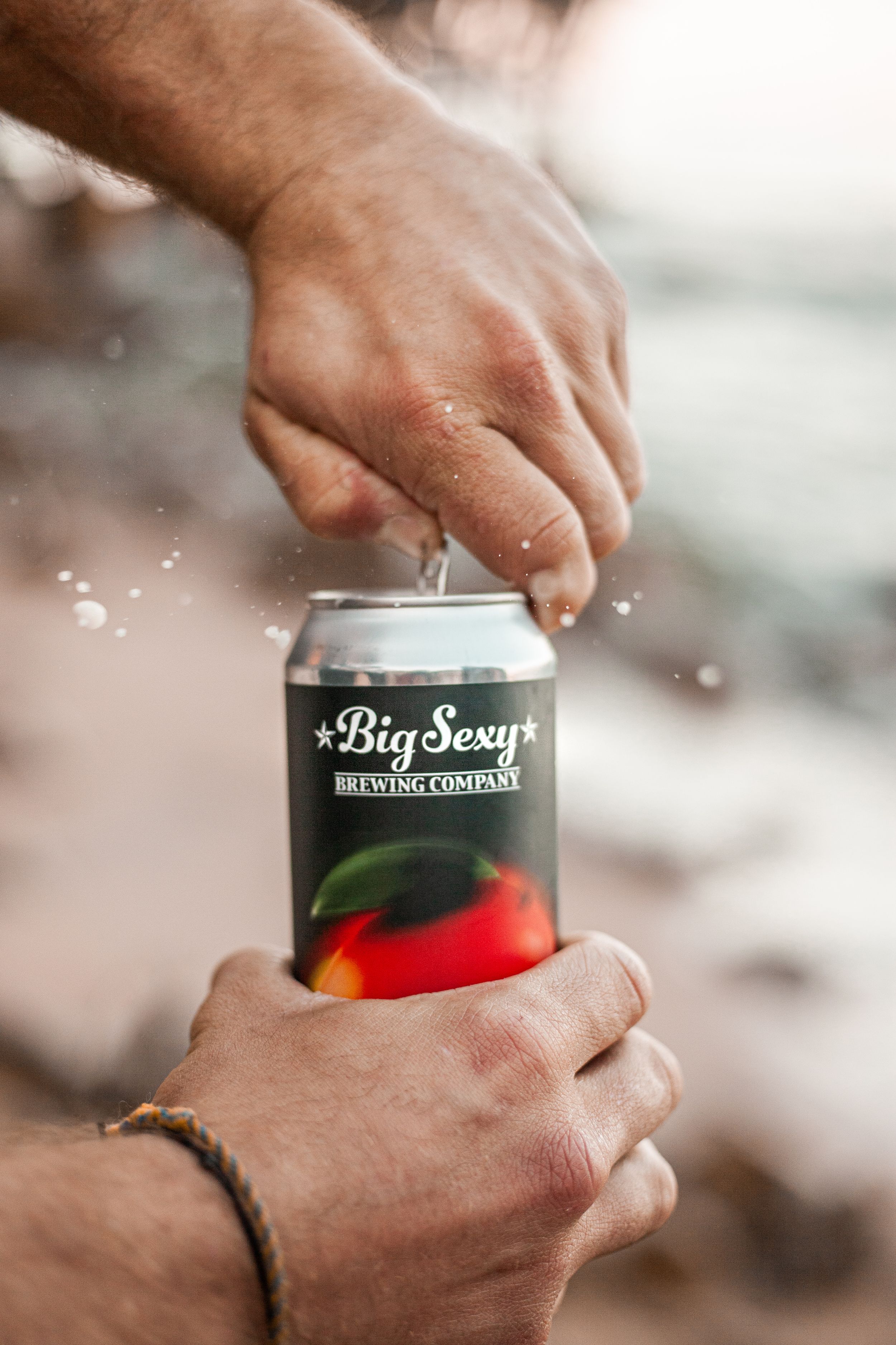 Hands opening a black can of Big Sexy branded beverage with water drops splashing.