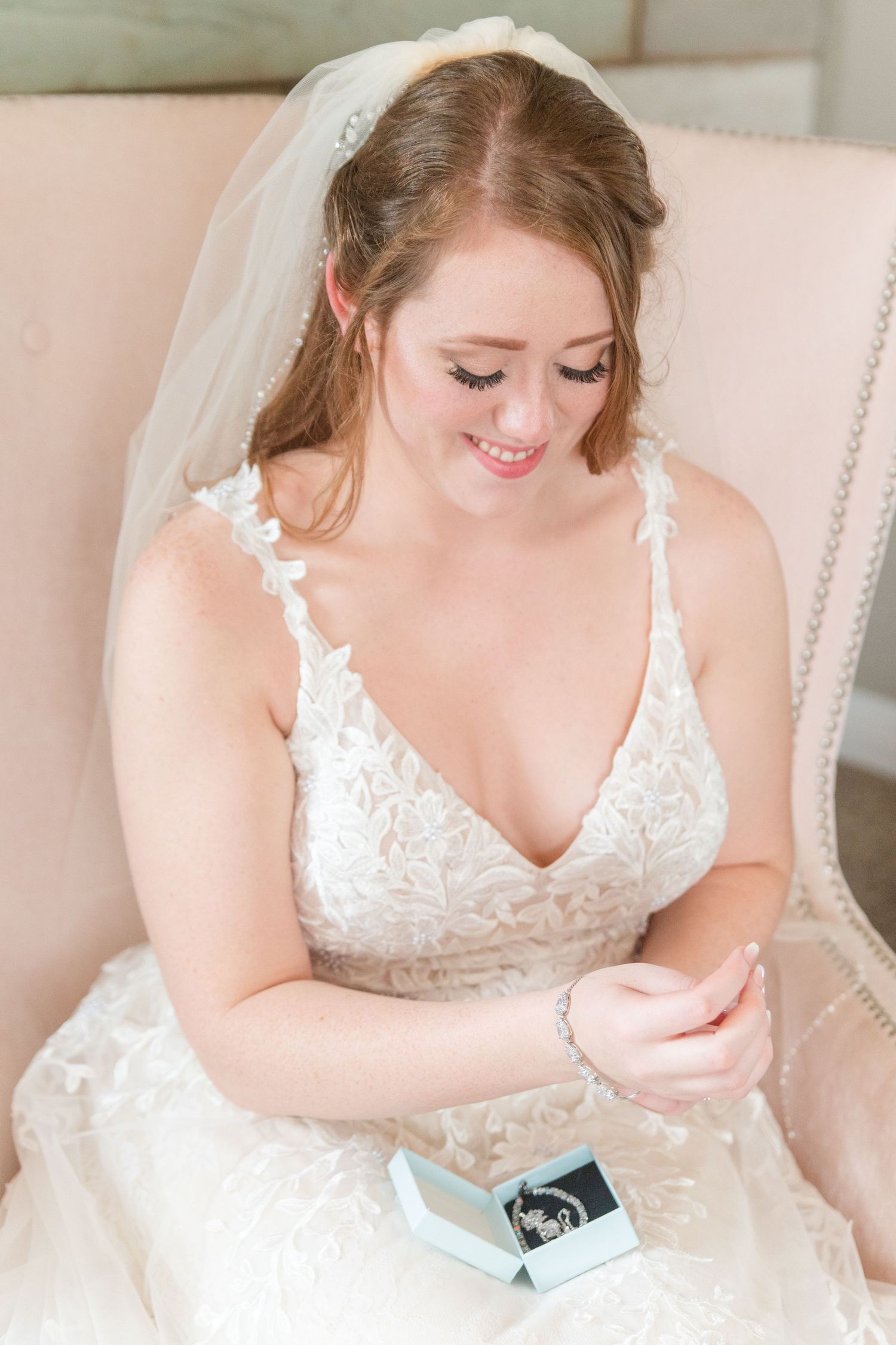 A person in a white lace wedding dress sits on a pink chair looking at something in their hands.