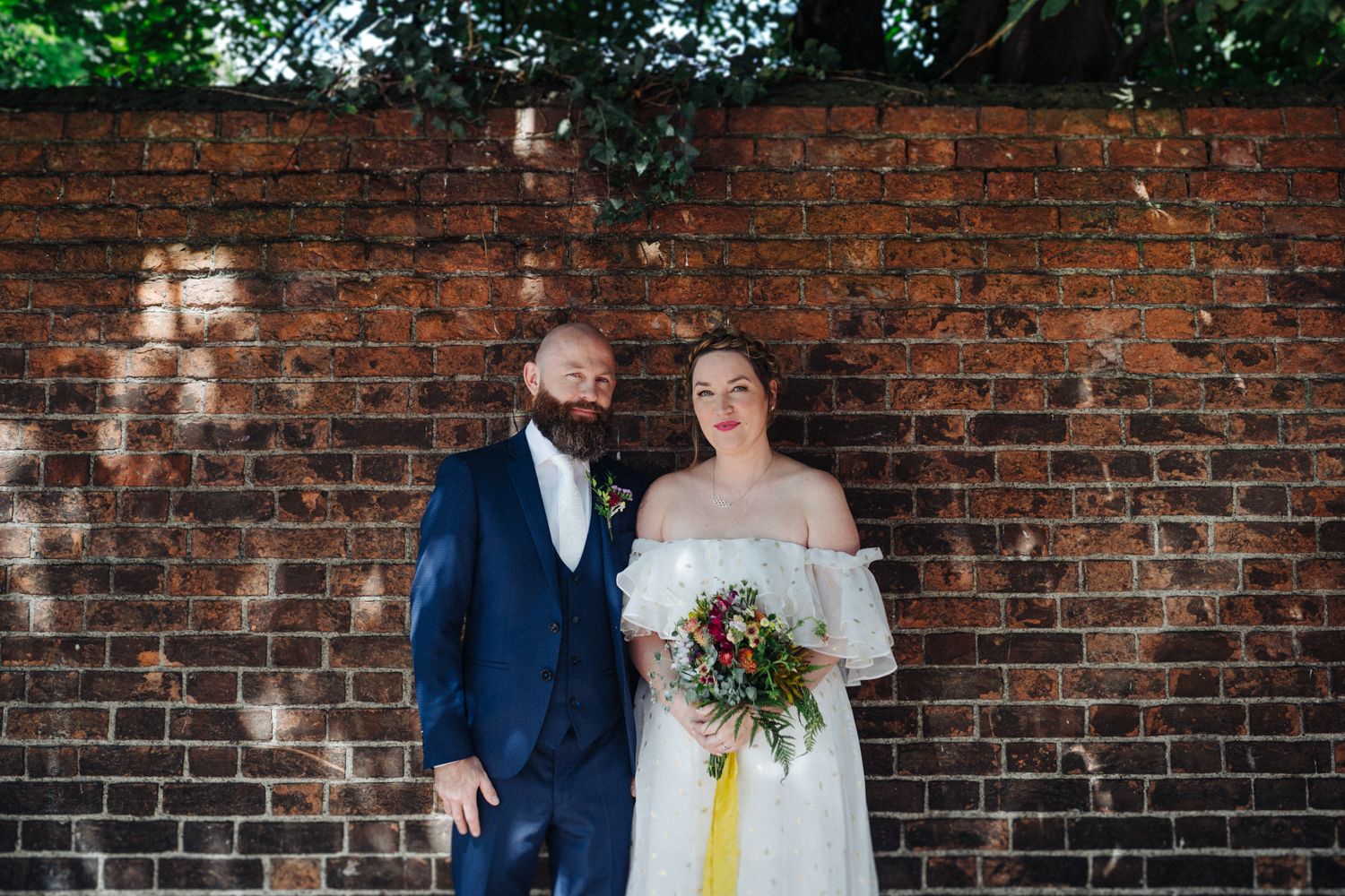 A couple stands against a brick wall during their wedding, with colorful florals.