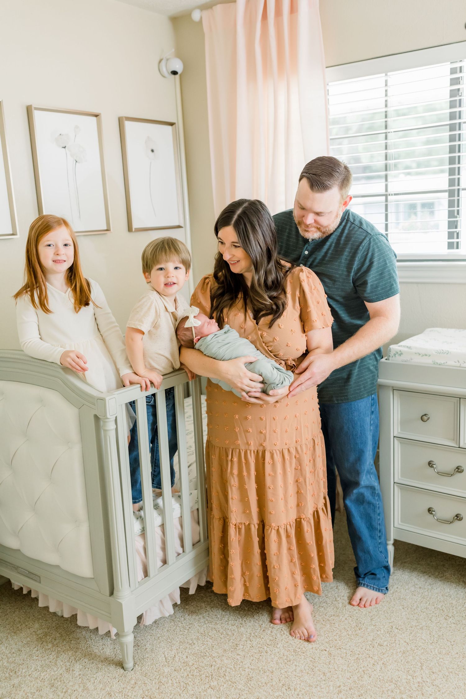 A family gathers around a gray crib in a nursery decorated with pink curtains and minimalist wall art.