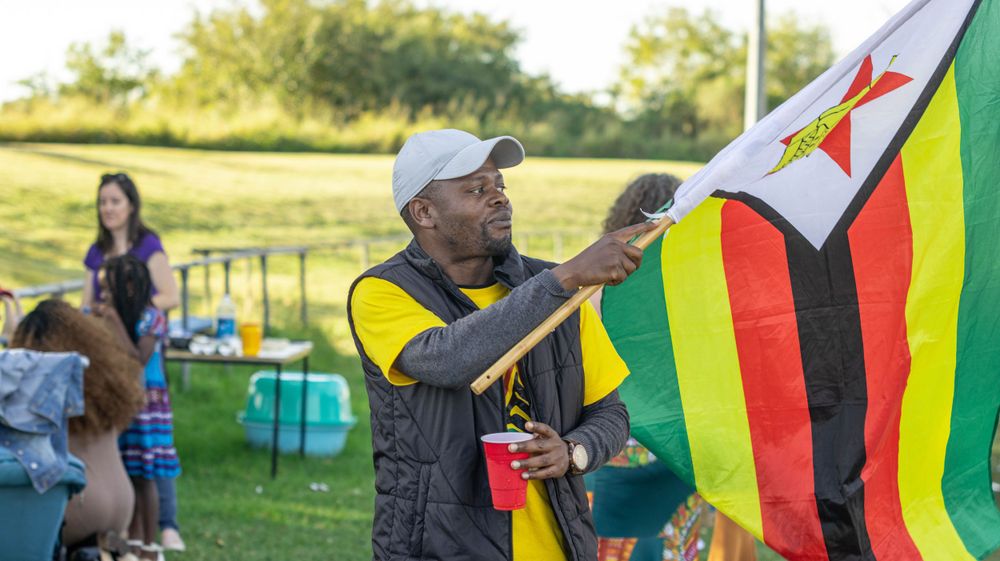 Person displaying Zimbabwe flag at outdoor festival with tents and people in background.