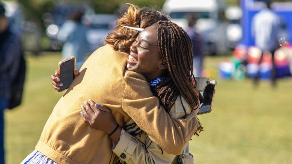 Two people sharing a warm embrace on a sunny grassy field during an outdoor event.