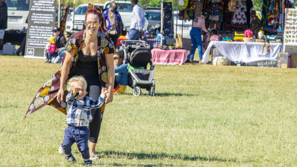 A young child runs on grass while others enjoy a community festival with vendor booths in background.