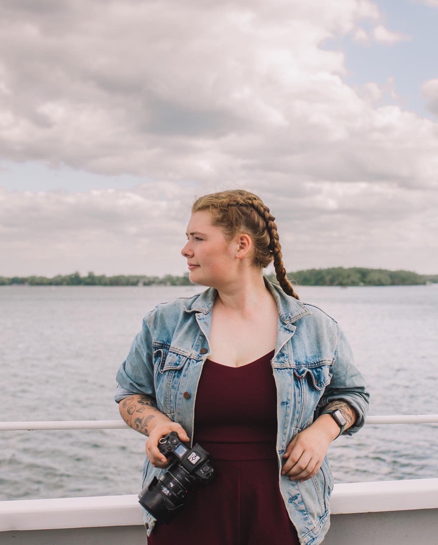 Photographer wearing denim jacket poses with camera against waterfront backdrop.