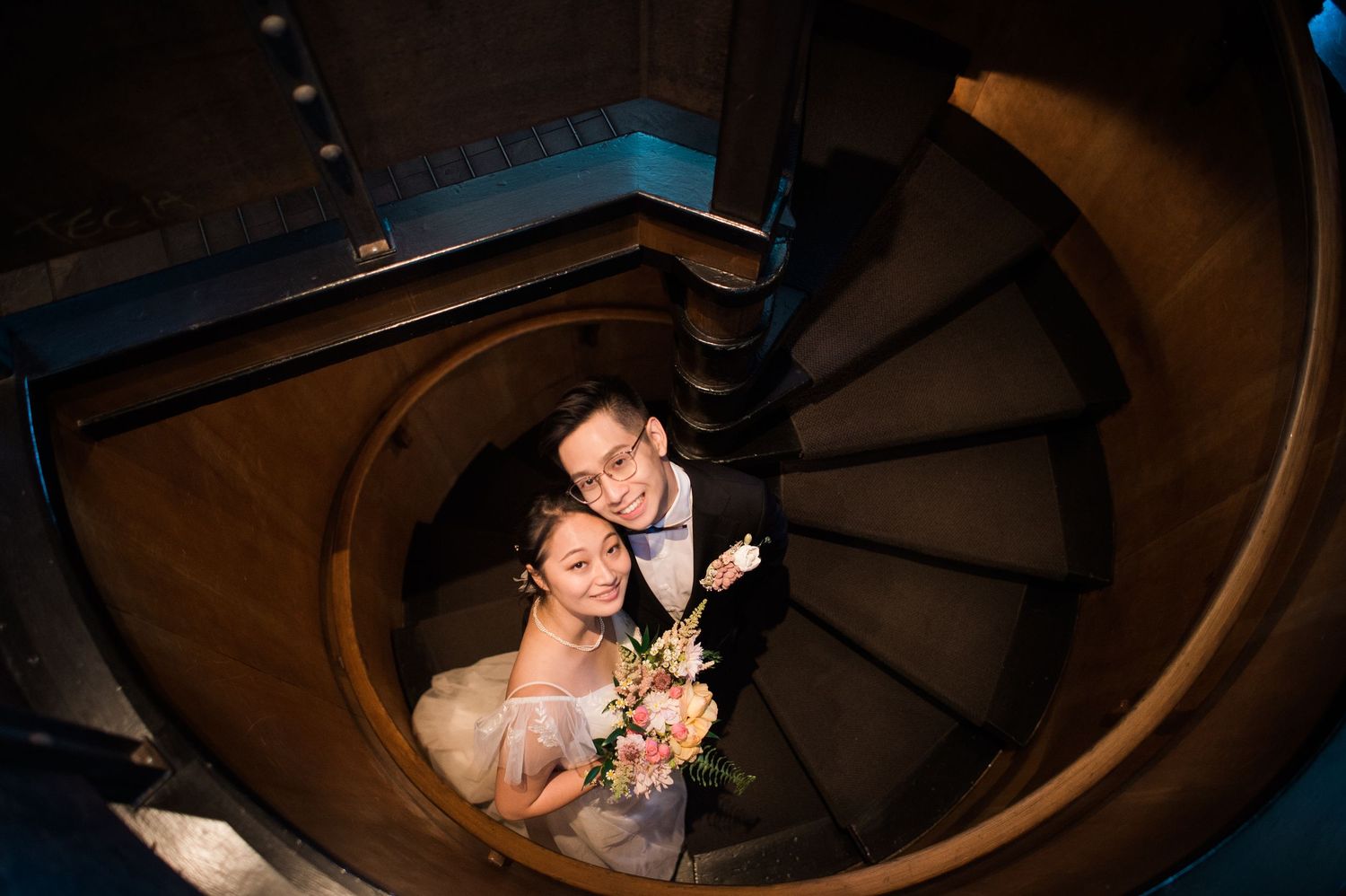 A romantic couple poses on a spiral staircase in dramatic lighting.