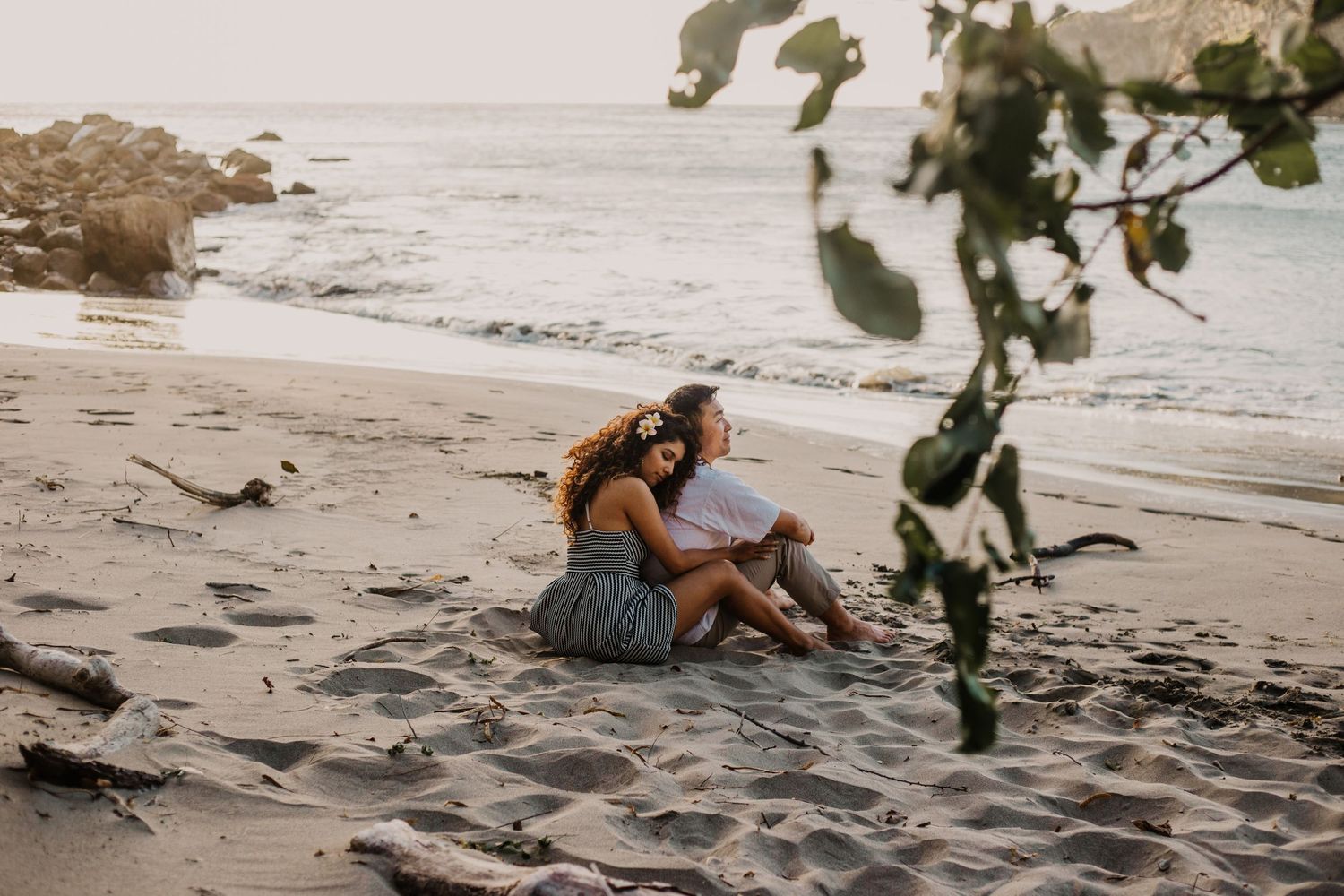 woman hugs husband from behind while sitting on the beach