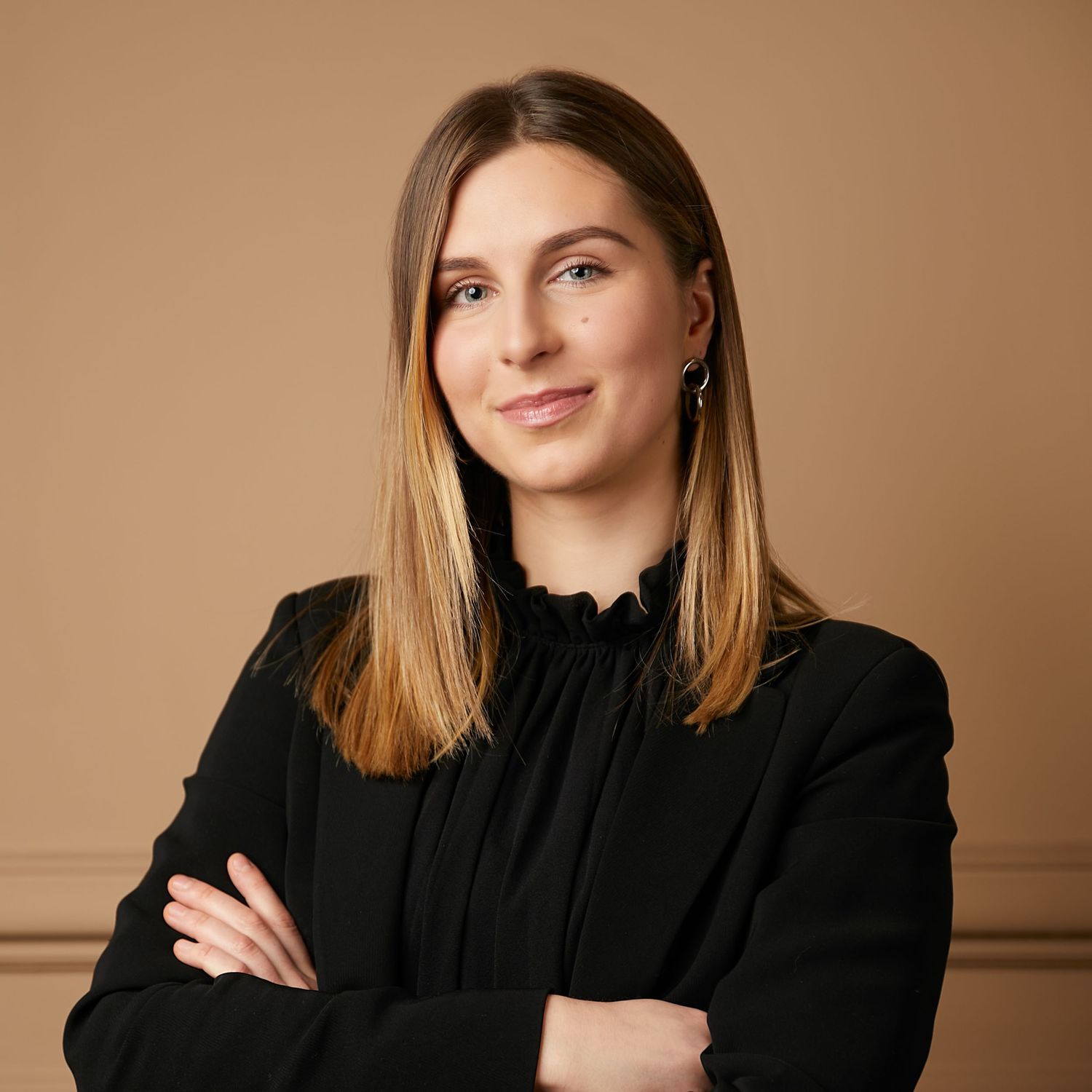 Professional studio portrait of someone in a black high-neck blouse with blue earrings against a brown background.
