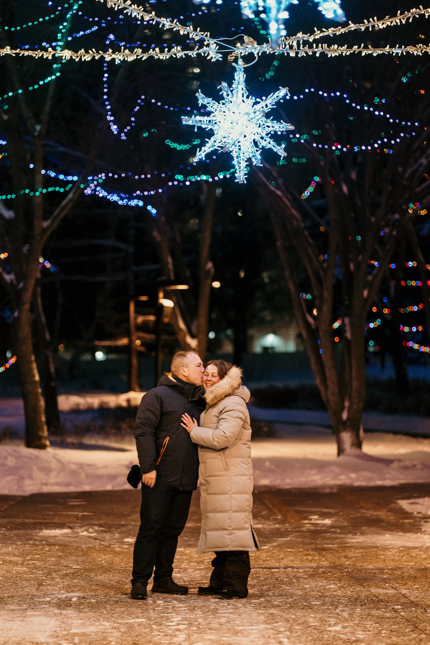 Winter scene with couple embracing under decorative snowflake lights and twinkling string lights.