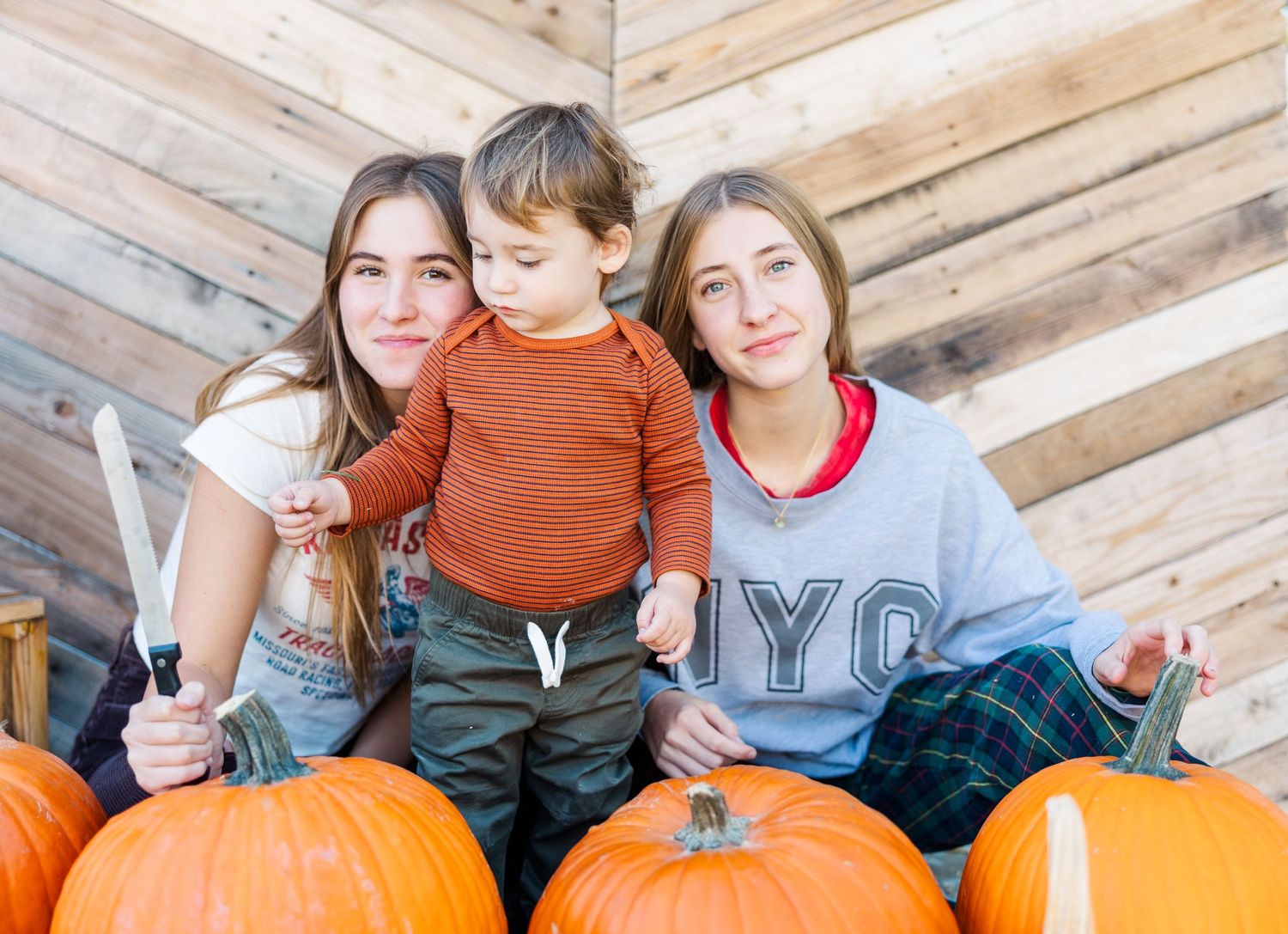 Group enjoying autumn festivities while seated among large orange pumpkins against wooden wall.