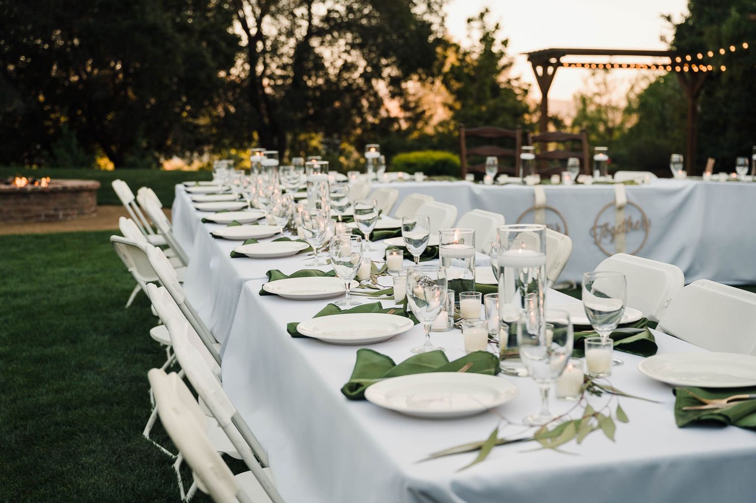 Long white dining table set for an elegant outdoor event with place settings and wine glasses in a garden setting.