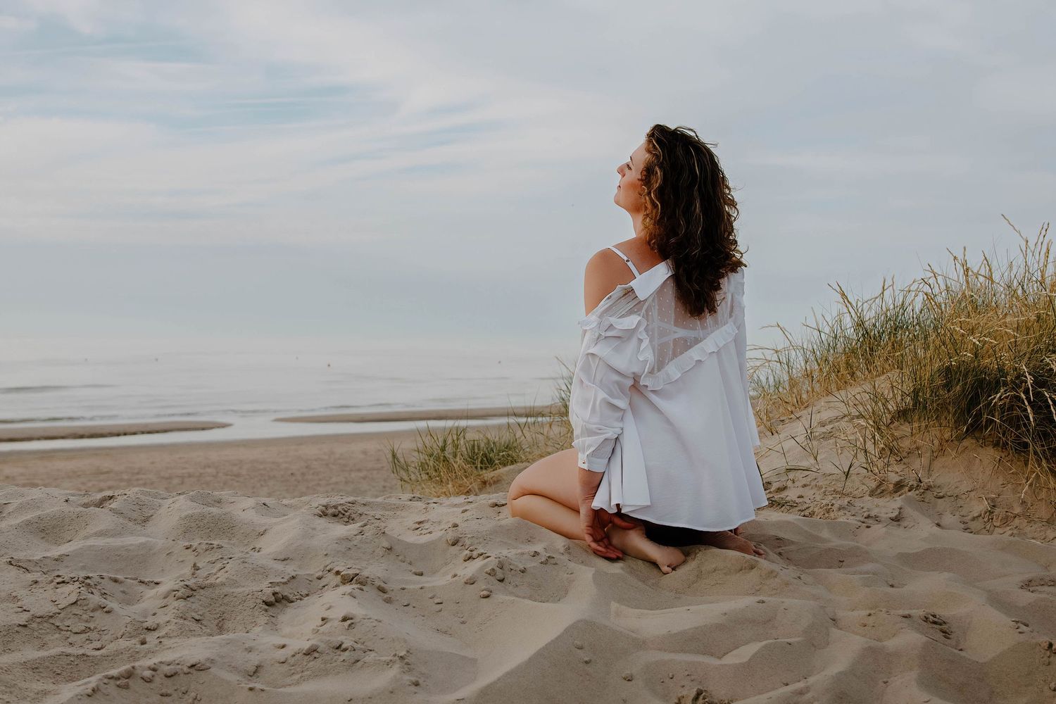Person in white shirt sits peacefully on sandy beach dunes overlooking ocean.