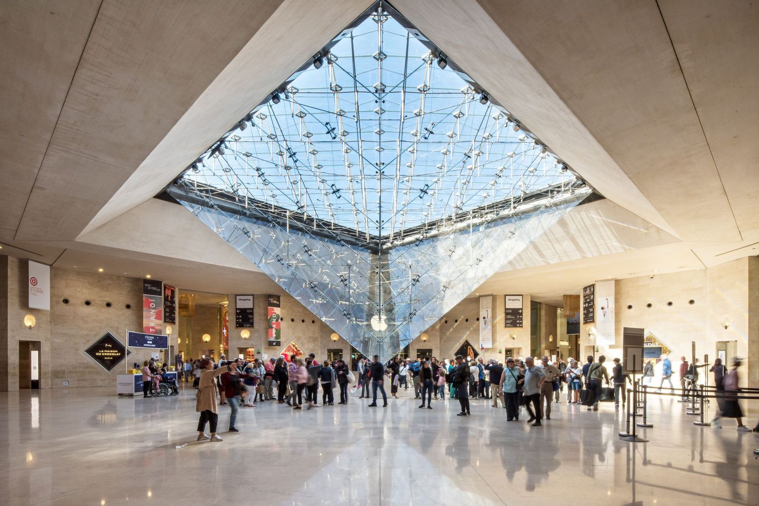 Crowded museum atrium with inverted glass pyramid skylight and marble floors.
