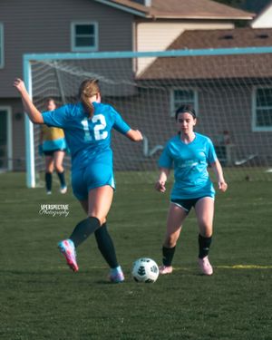 Two soccer players in bright blue uniforms compete for the ball on a grass field with goal nets visible.