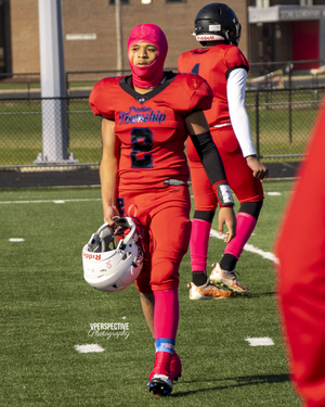 Football player in all red uniform with pink accents walks on field carrying helmet.