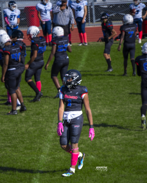 Football player wearing pink gloves stands on field during game with teammates in background.