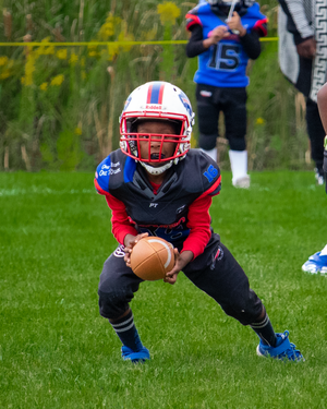 Youth football player in red and blue uniform crouches in ready position with ball.