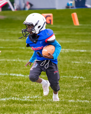 Youth athlete in blue and white uniform carries football while running on grass field.