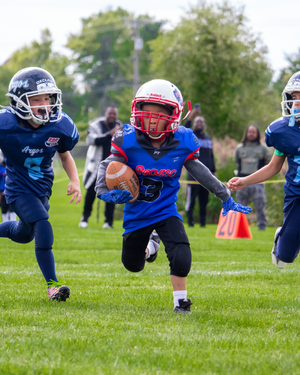 Young football player in blue and red uniform runs with the ball between two defenders during a youth game.